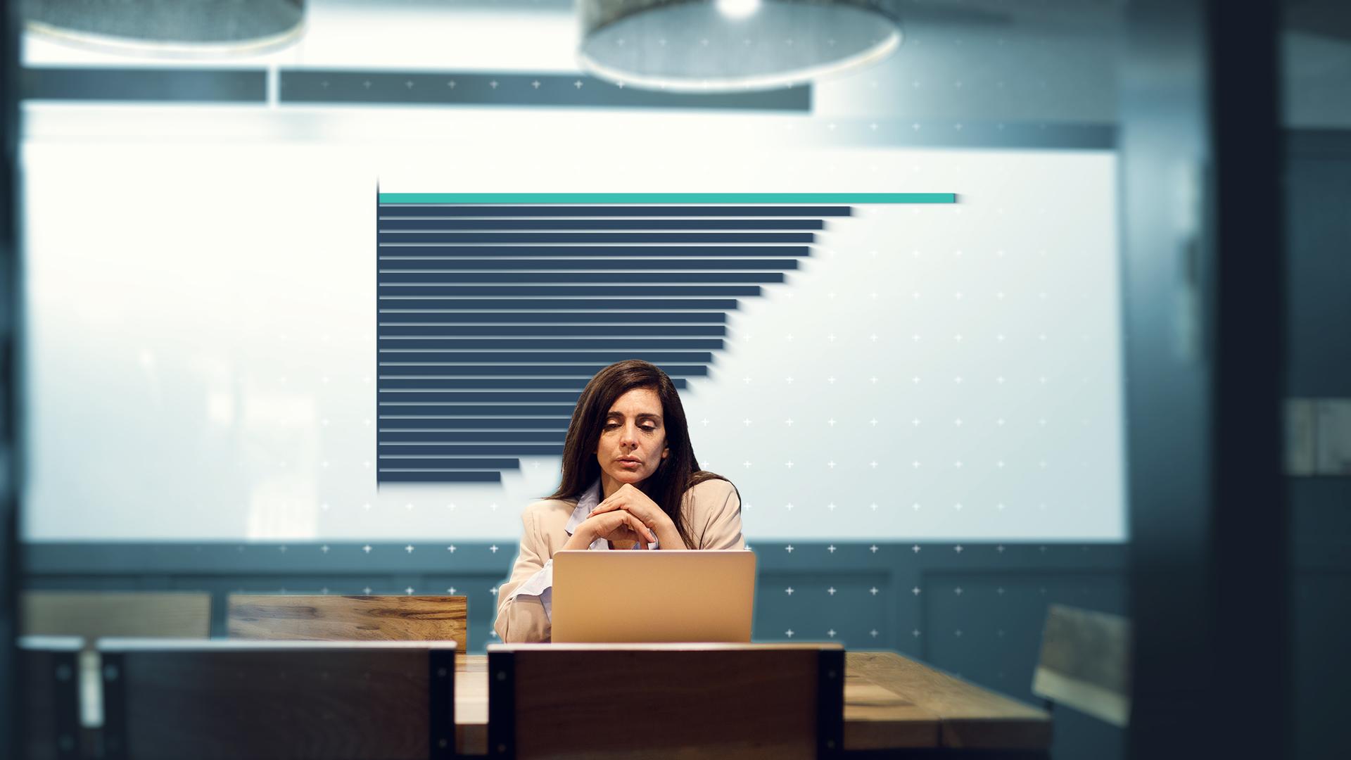 Eine Frau sitzt vor einem Laptop und im Hintergrund ist ein Balkendiagramm auf einem Whiteboard zu sehen.