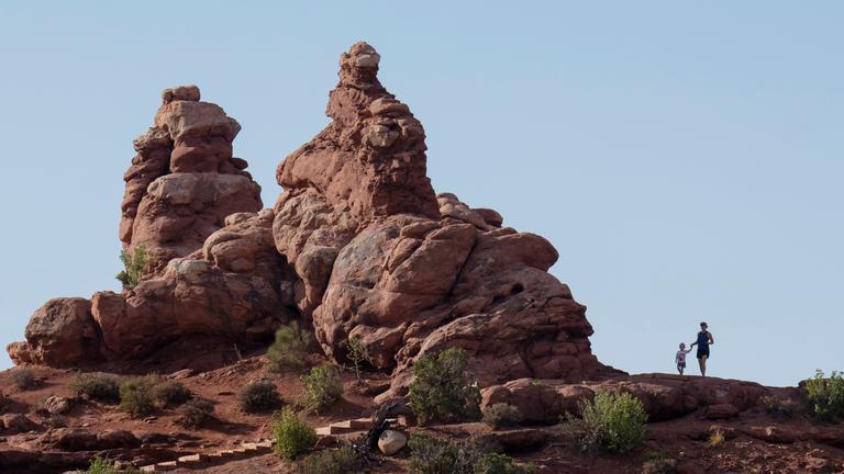 Zwei Menschen stehen neben einer roten Felsformation in der Wüste.