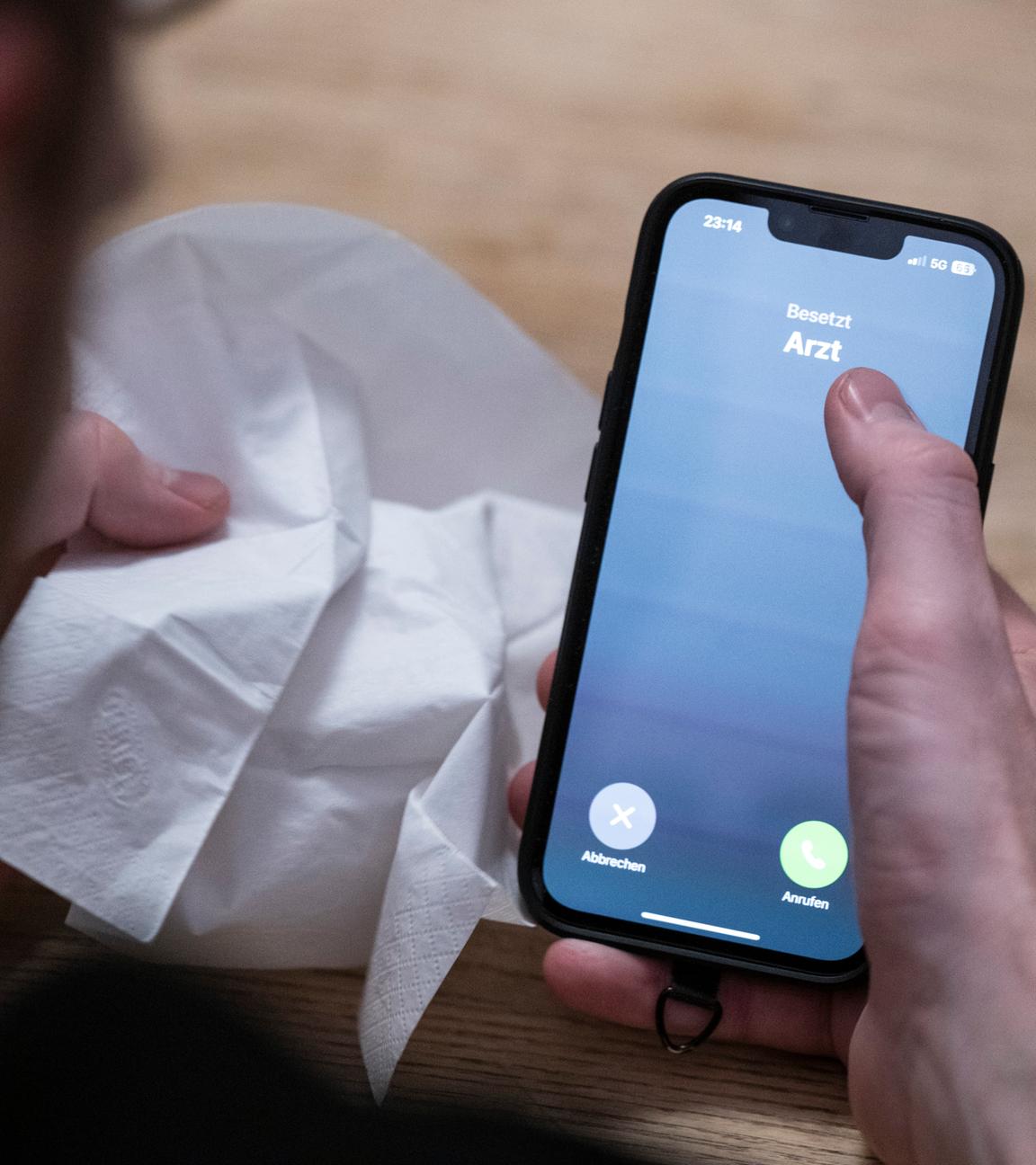Ein Mann hält ein Taschentuch in einer Hand und ein Telefon mit dem Schriftzug 'Arzt' in der Anderen.