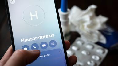 Symbolbild: Eine Person hält ein Smartphone in der Hand und ruft eine Hausarztpraxis an. 
