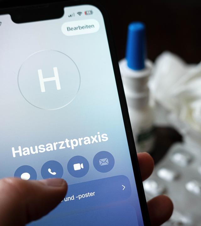 Symbolbild: Eine Person hält ein Smartphone in der Hand und ruft eine Hausarztpraxis an. 