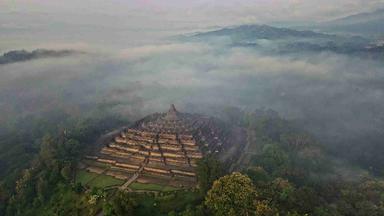 Ein alter Tempel aus Stein mit Stufenpyramide steht in einem Wald und ist von Nebel und hügeliger Landschaft umgeben.