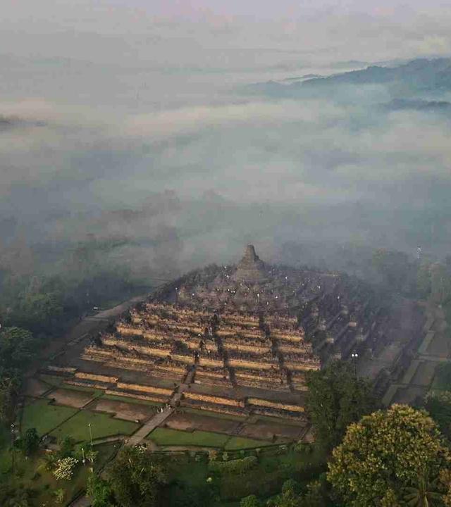 Ein alter Tempel aus Stein mit Stufenpyramide steht in einem Wald und ist von Nebel und hügeliger Landschaft umgeben.