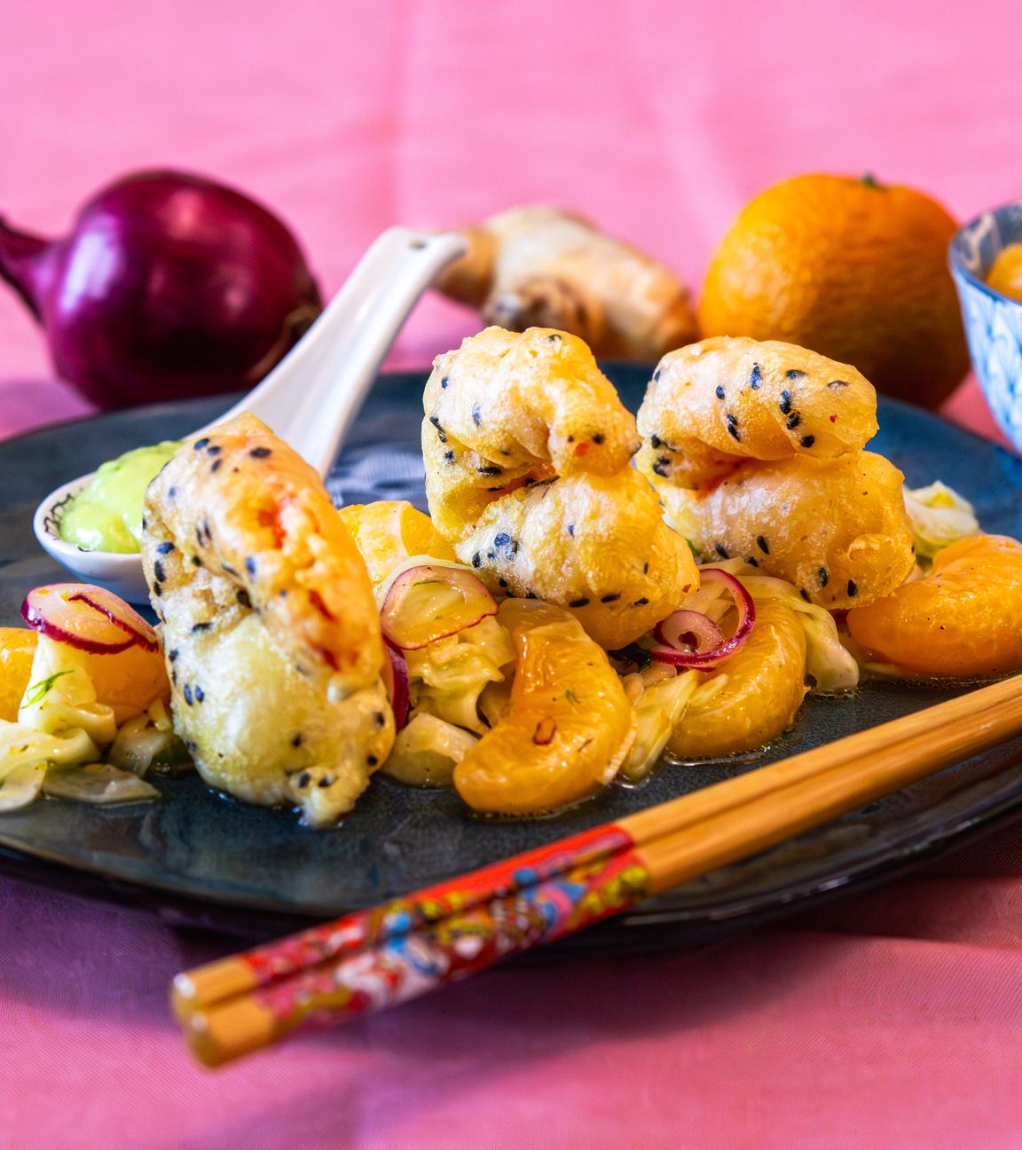 Auf einem schwarzen Teller ist das Gericht Tempura-Garnelen mit Mandarinen-Fenchel-Salat. Im Hintergrund liegen geschälte Mandarinen, eine Zwiebel und eine Knolle Ingwer.