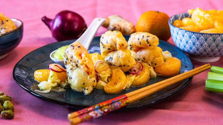 Auf einem schwarzen Teller ist das Gericht Tempura-Garnelen mit Mandarinen-Fenchel-Salat. Im Hintergrund liegen geschälte Mandarinen, eine Zwiebel und eine Knolle Ingwer.