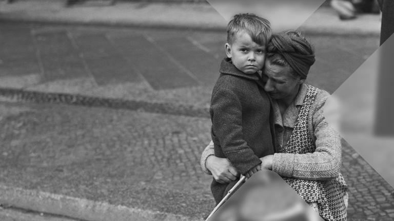 Rechts im Bild sitzt eine Frau auf der Bordsteinkante. Sie hält einen kleinen Jungen in ihrem Armen und weint. Der Junge schaut traurig in die Kamera. 