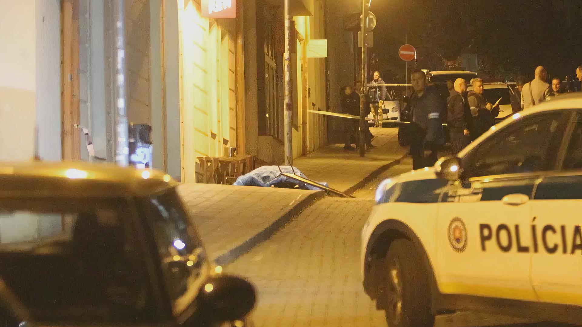 Nächtliche innerstädtische Straßenszene: Vor einer Häuserfront im gelben Licht der Straßenlaternen liegt ein mit Folie zugedecktes Todesopfer auf Gehweg. Davor parkt ein slowakisches Polizeiauto, Polizisten stehen daneben.