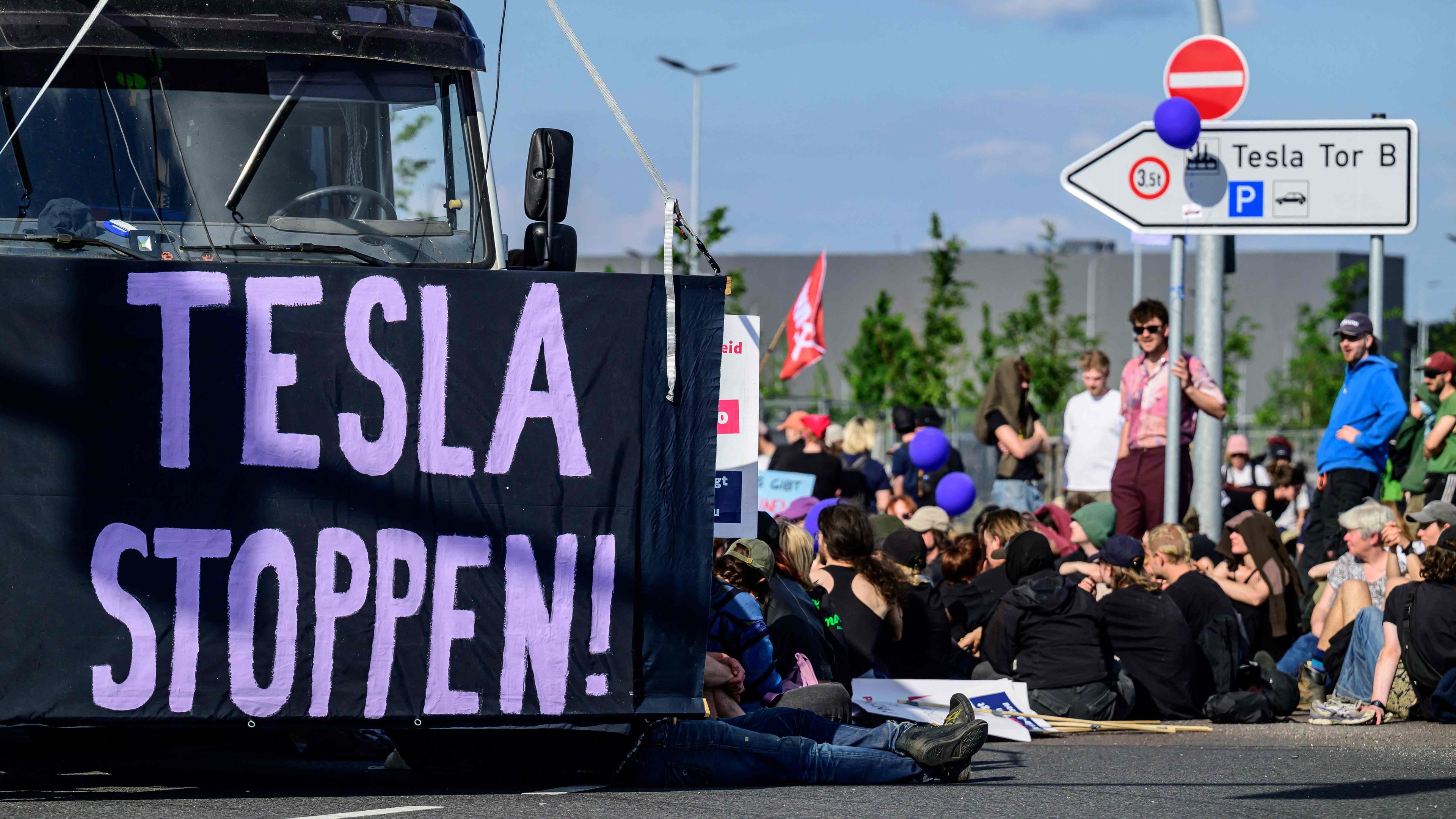 Klimaaktivisten sitzen auf dem Boden und blockieren das Tesla Gelände in Grünheide. Links ist ein Banner der Aufschrift "TESLA STOPPEN" vor einen LKW gespannt.