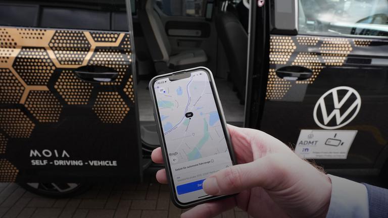 Hamburg: Ein Mitarbeiter hält ein Smartphone mit der Moia-App vor einen autonom fahrenden ID. Buzz AD von Volkswagen (gestellte Szene).
