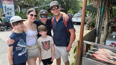 Eltern stehen mit ihren drei Kindern auf einer Straße in Thailand. Alle schauen lächelnd in die Kamera. Neben ihnen liegen Fische auf einem Grill, im Hintergrund befinden sich Autos und ein Motorrad.