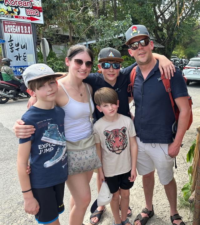 Eltern stehen mit ihren drei Kindern auf einer Straße in Thailand. Alle schauen lächelnd in die Kamera. Neben ihnen liegen Fische auf einem Grill, im Hintergrund befinden sich Autos und ein Motorrad.