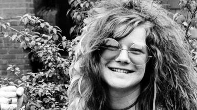  Schwarz-Weiß-Foto von Janis Joplin. Sie trägt eine große runde Brille und grinst mit wilder Haar-Mähne in die Kamera.