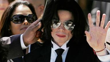 Michael Jackson mit Anzug und Krawatte inmitten einer Menschengruppe. Eine Hand hält ihm von links einen Sonnenschirm über den Kopf. Jackson hat die linke Hand erhoben.