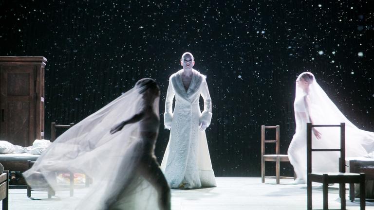 Auf einer Bühne mit weißem Boden und schwarzem weiß gesprenkelten Hintergrund laufen im Vordergrund zwei mit großen durchsichtigen Schleiern verhangene Frauen aufeinander zu, während in der Mitte eine Person in langem weißen Mantel steht.