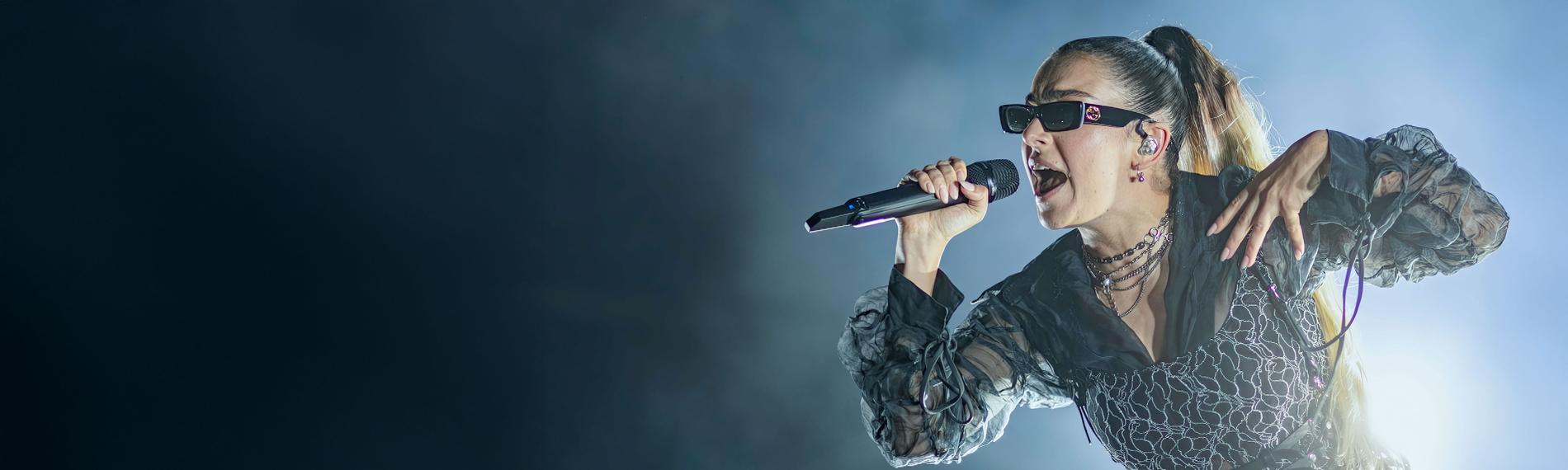 Charli xcx trägt eine Sonnenbrille und singt in ein Mikrofon. Der Hintergrund ist dunkel, auf einer Seite ist Nebel.