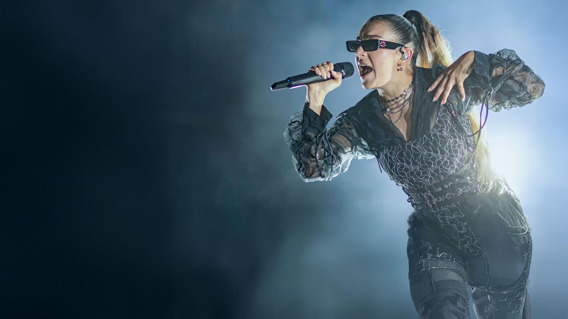 Charli xcx trägt eine Sonnenbrille und singt in ein Mikrofon. Der Hintergrund ist dunkel, auf einer Seite ist Nebel.
