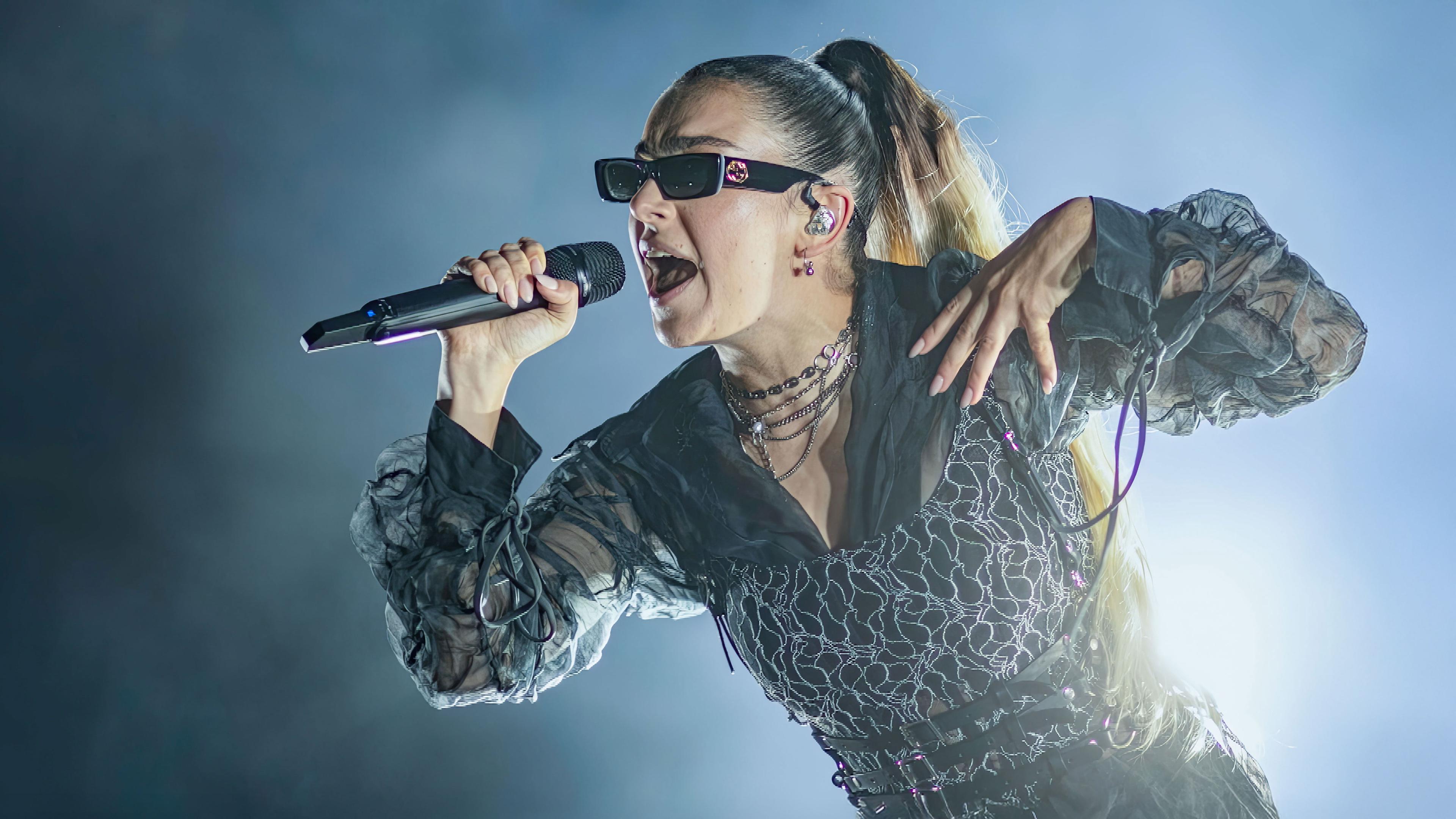 Charli xcx trägt eine Sonnenbrille und singt in ein Mikrofon. Der Hintergrund ist dunkel, auf einer Seite ist Nebel.