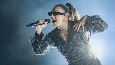 Charli xcx trägt eine Sonnenbrille und singt in ein Mikrofon. Der Hintergrund ist dunkel, auf einer Seite ist Nebel.