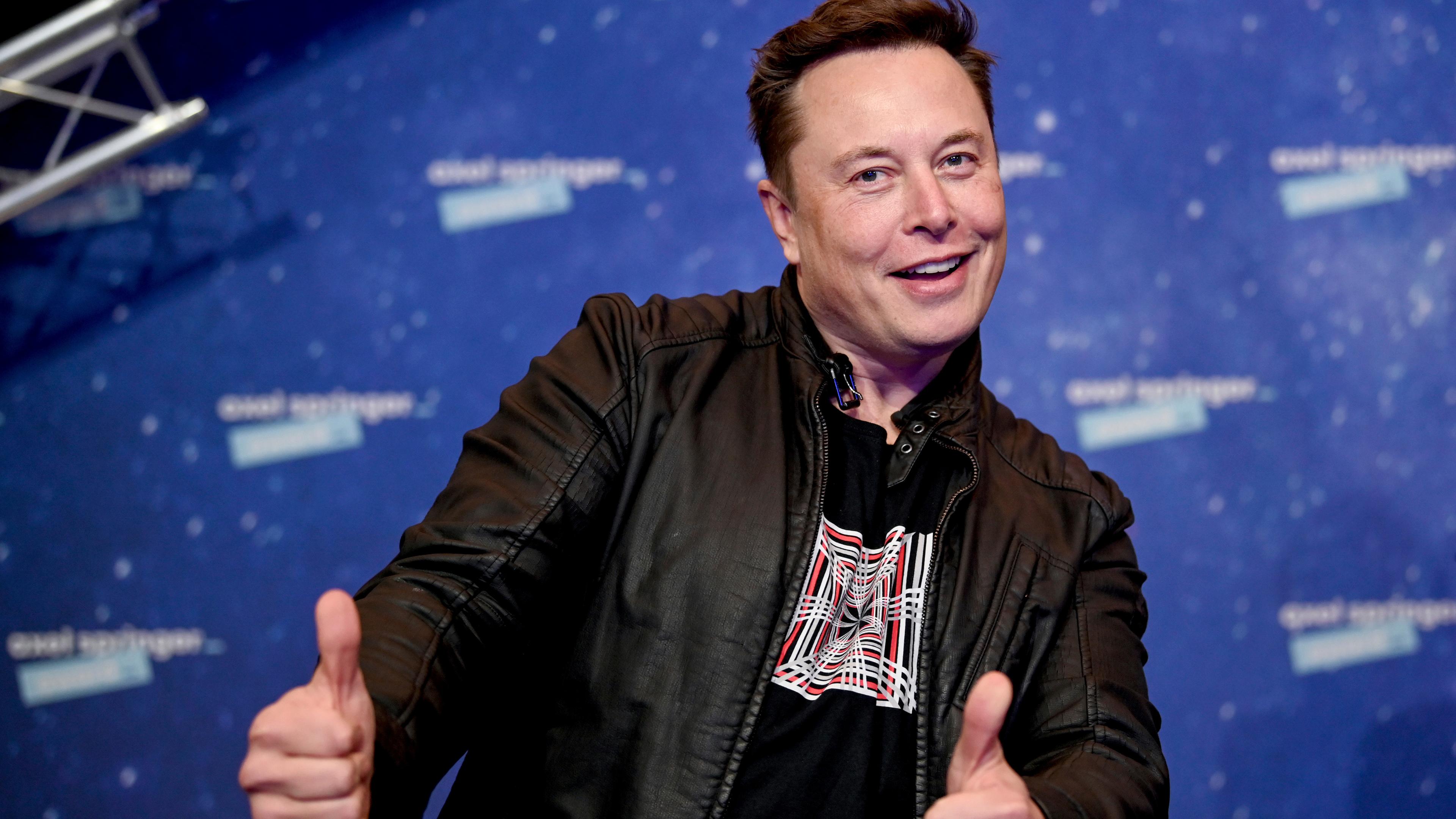 Elon Musk steht vor einer blauen Wand und zeigt zwei Daumen hoch in die Kamera.