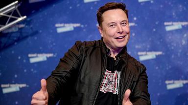 Elon Musk steht vor einer blauen Wand und zeigt zwei Daumen hoch in die Kamera.