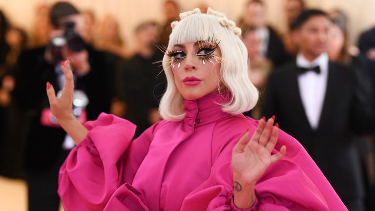  Lady Gaga trägt ein rosafarbenes Kleid mit Puffärmeln. Hellblonde Haare reichen ihr bis zur Schulter und sie trägt lange künstliche Wimpern. Im Hintergrund sind Fotografen und ein Mann im Smoking zu sehen.