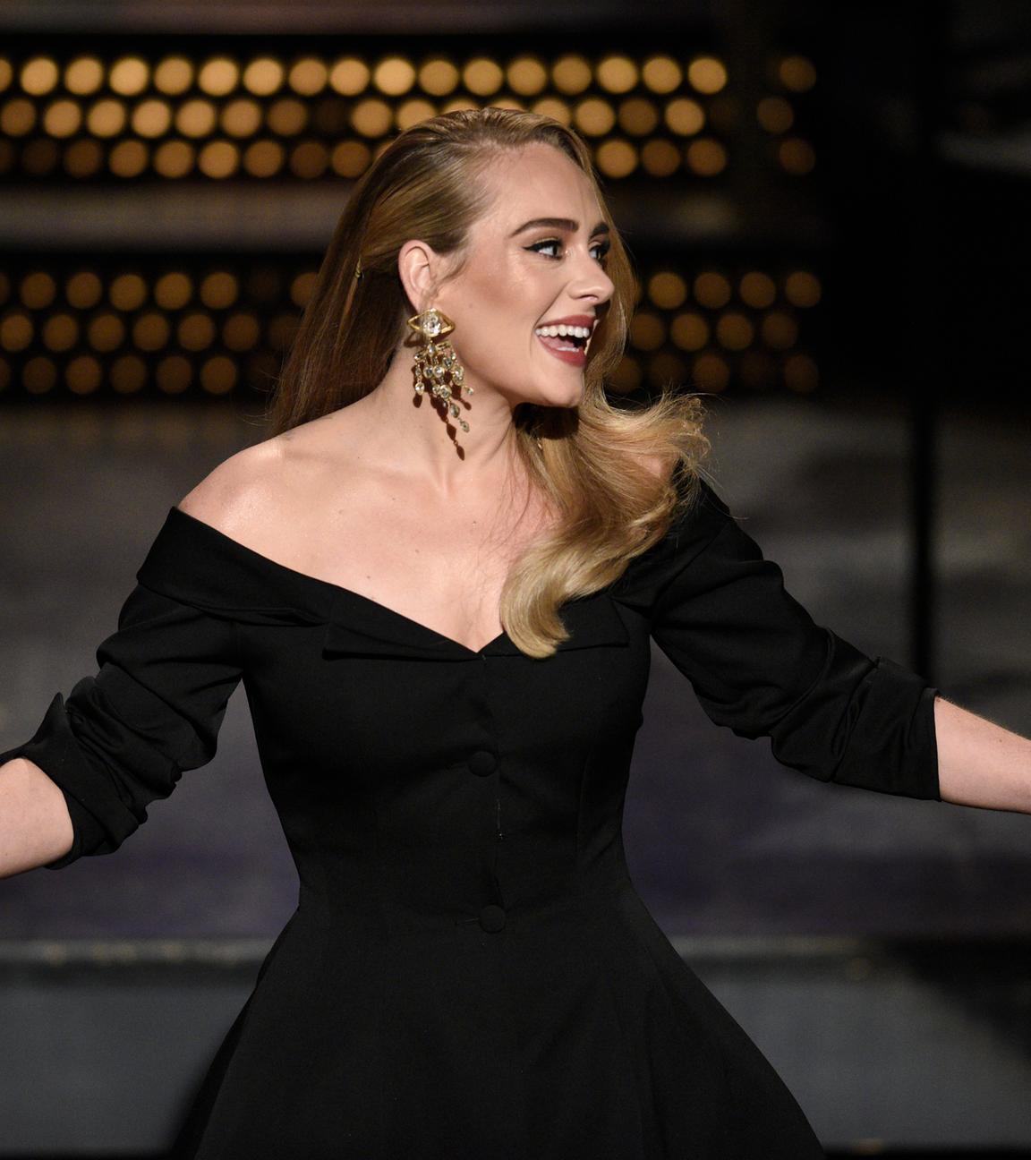 Adele mit offenem Haar, einem schwarzen Kleid und großen Goldohrringen steht auf einer Bühne, schaut nach rechts und lächelt. Sie streckt die Arme aus.