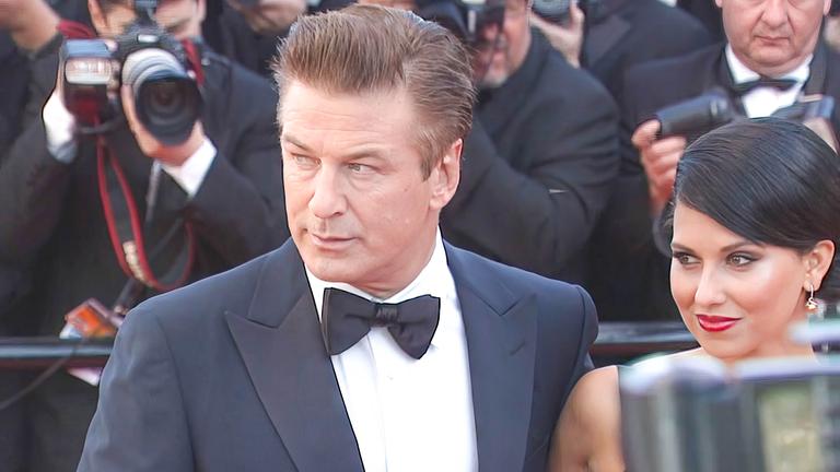 Alex Baldwin steht in Anzug und Fliege neben seiner dunkelhaarigen Frau. Beide schauen zur Seite, hinter ihnen stehen Fotografen.