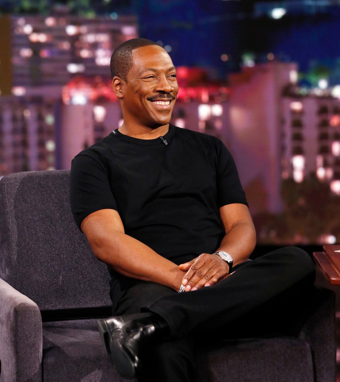 Eddie Murphy sitzt schwarz gekleidet auf einem Sessel in einer Show und lacht.