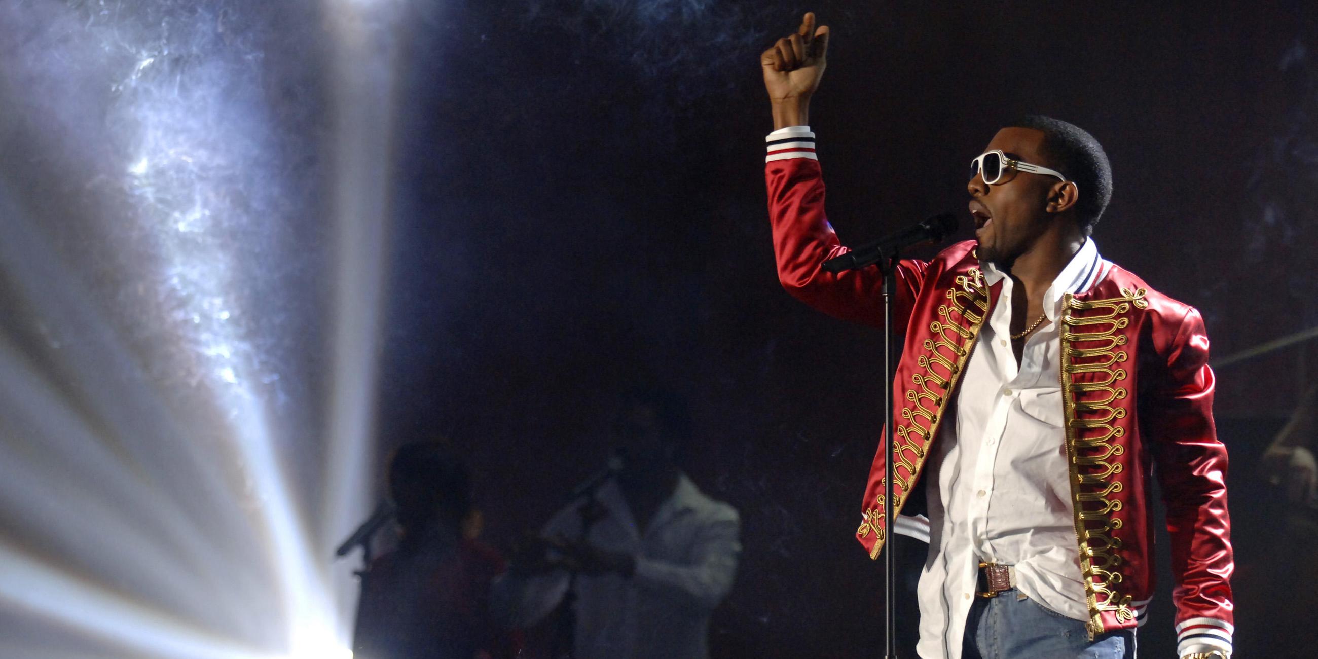 Rapper Kanye West steht auf der Bühne vor einem Mikrofon und singt. Die Bühne ist dunkel, an der Seite leuchtet grelles Scheinwerferlicht.