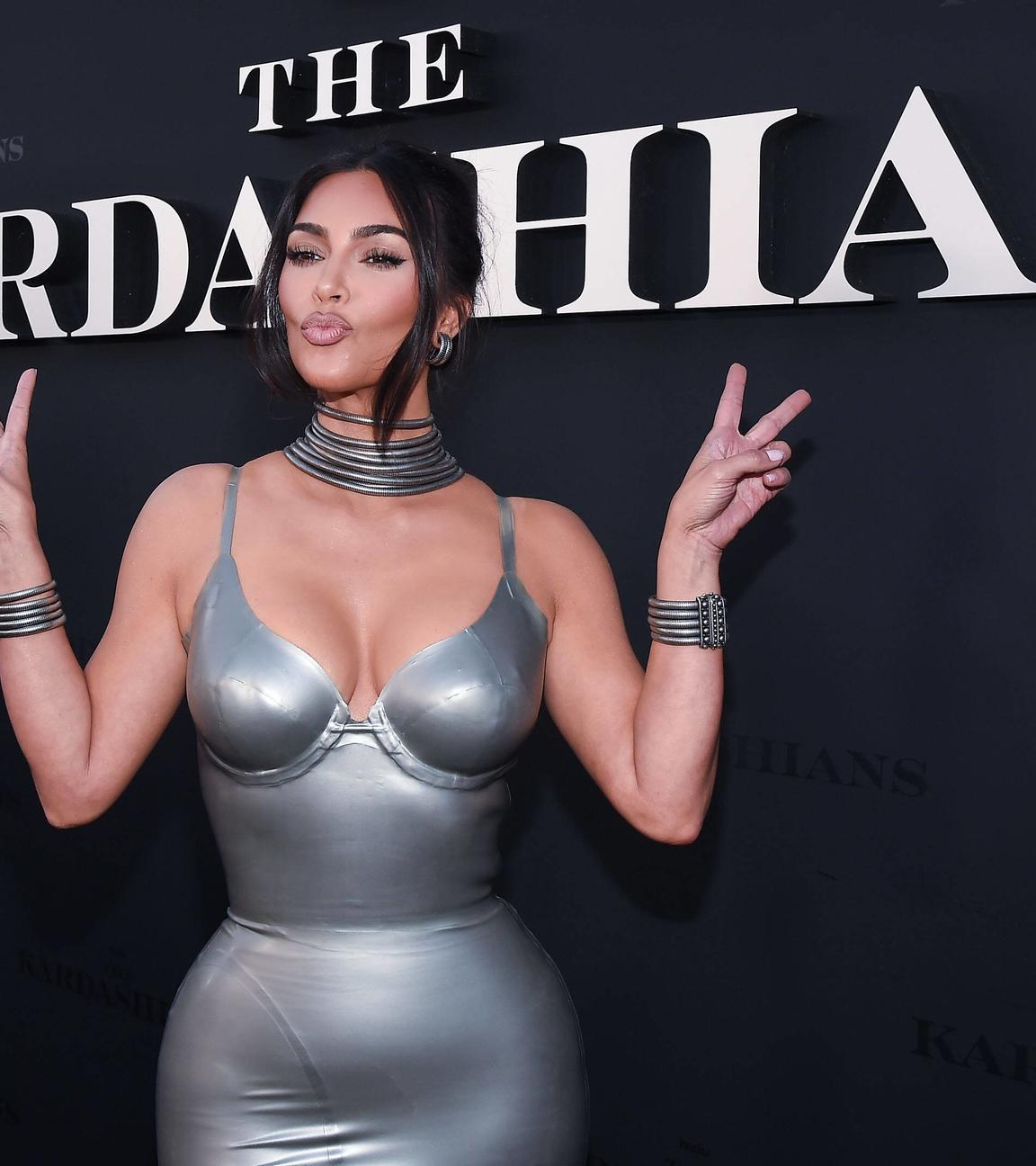 Kim Kardashian steht vor einer schwarzen Wand mit der Aufschrift "The Kardashians". Sie trägt ein silberfarbenes Kleid und silbernen Schmuck. Sie macht einen Kussmund und zeigt mit beiden Händen das Peace-Zeichen.