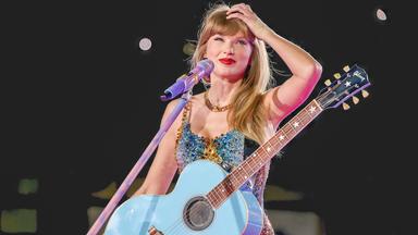 Taylor Swift steht mit Gitarre an einem Mikrofon. Sie lächelt und fasst sich an den Kopf.
