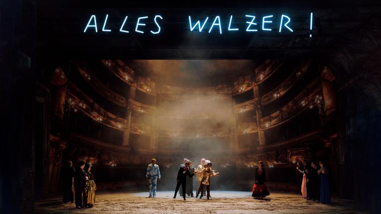 In einer Leuchtschrift und in Großbuchtsaben steht oben im Bild: "Alles Walzer!". Im Hintergrund ist der Zuschauersaal der Wiener Volksoper zu erkennen und im Vordergrund unten ist die Bühne, auf der mehrere Personen in Anzügen und Paillettenkleidern stehen.