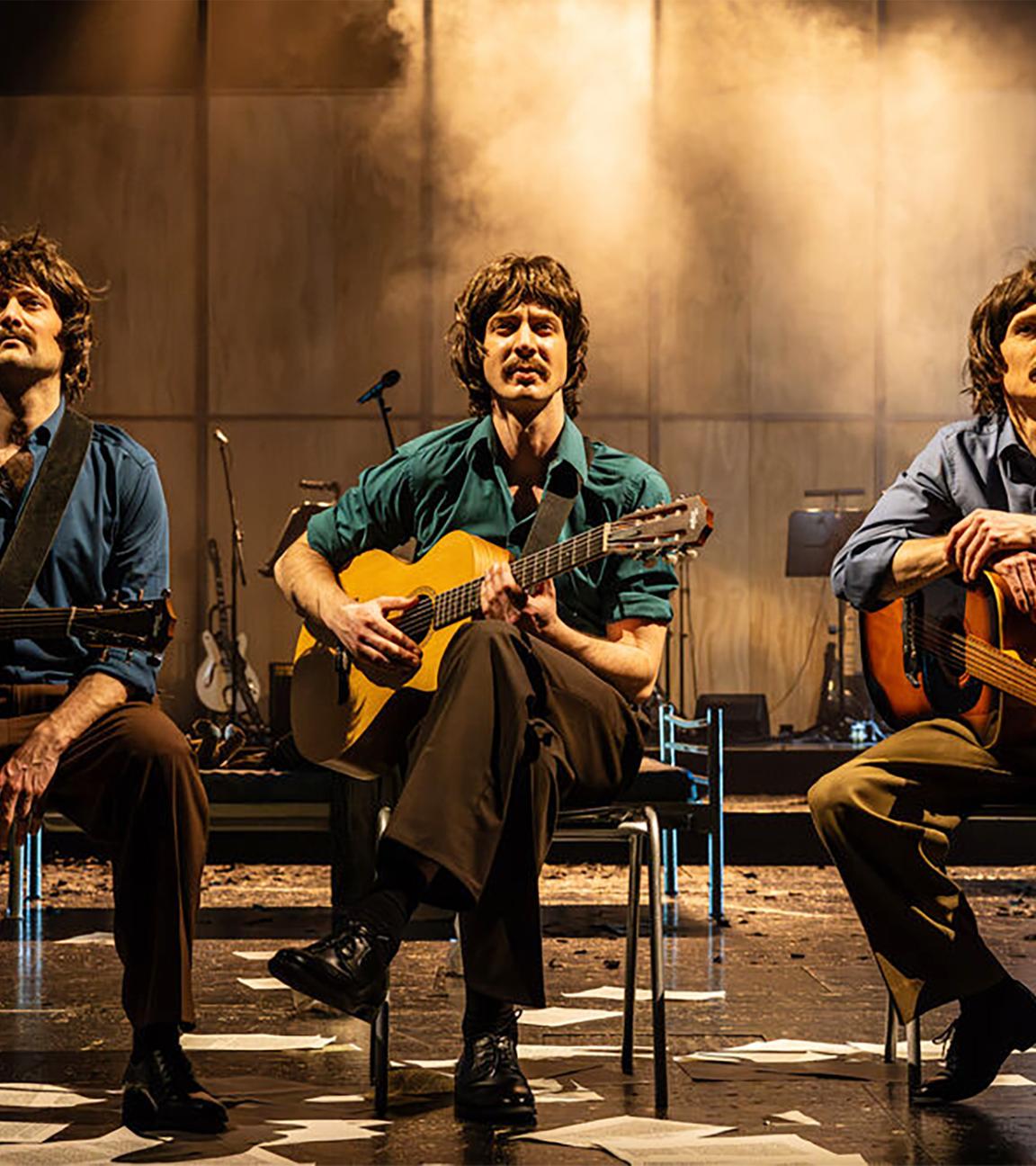 Bühnenszene: Drei Männer mit Gitarren, die Bekleidung der 1970er Jahre tragen, sitzen auf Stühlen.