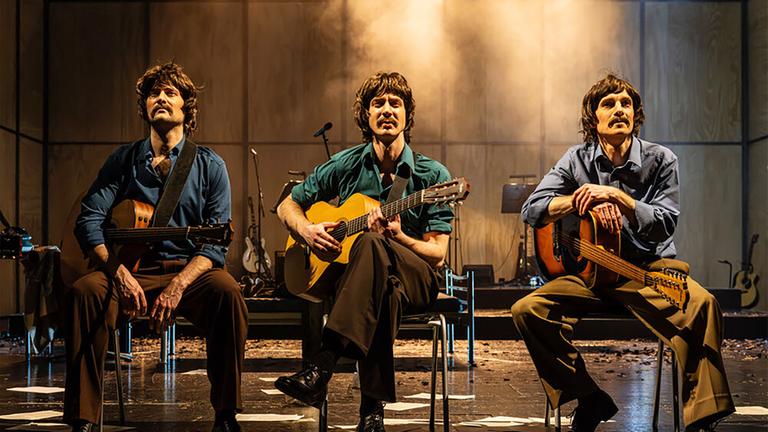Bühnenszene: Drei Männer mit Gitarren, die Bekleidung der 1970er Jahre tragen, sitzen auf Stühlen.