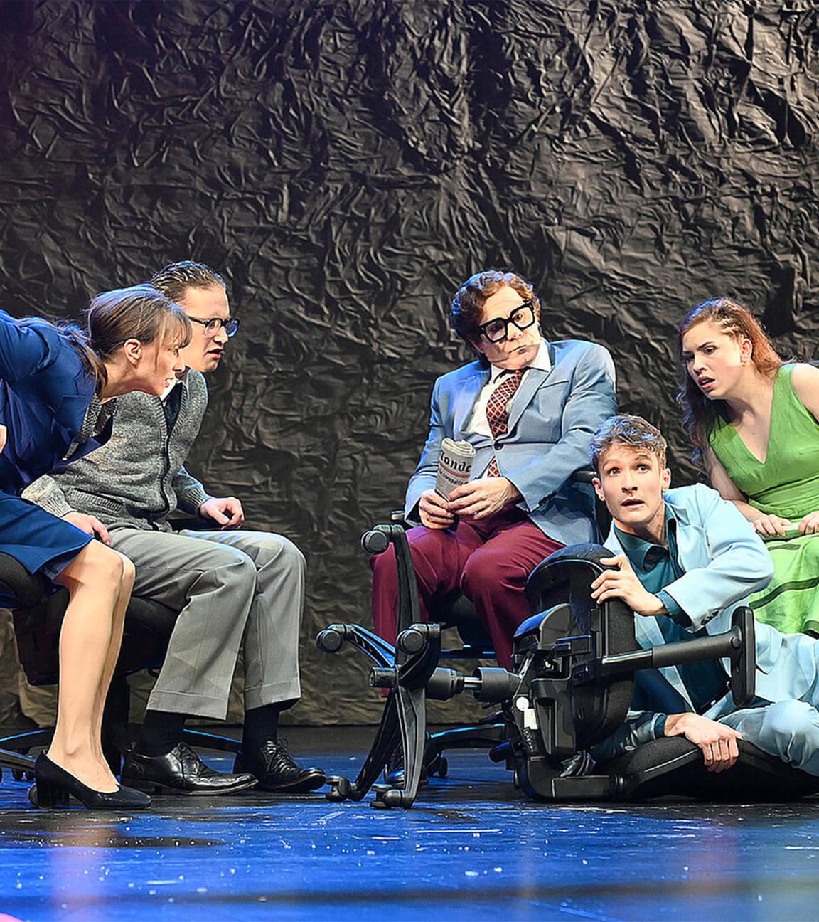 Bühnenszene: Mehrere Personen sitzen auf Bürostühlen im Halbkreis und schauen aufgeregt zu einem Mann, der auf dem Boden neben seinem umgefallenen Stuhl liegt.