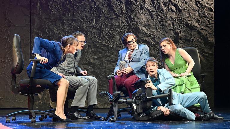 Bühnenszene: Mehrere Personen sitzen auf Bürostühlen im Halbkreis und schauen aufgeregt zu einem Mann, der auf dem Boden neben seinem umgefallenen Stuhl liegt.
