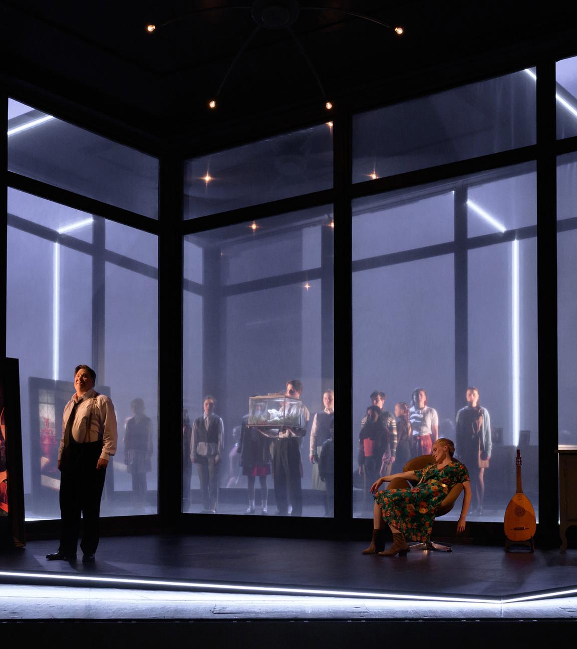 Bühnenszene: Man sieht die große Glasfensterfront eines Raumes im Halbdunkel. Links im Raum steht ein Mann neben einem Renaissancegemälde, rechts im Bild sitzt eine Frau in einem Sessel, neben einer Kommode. Draußen vor den Fenstern stehen Menschen und schauen hinein.