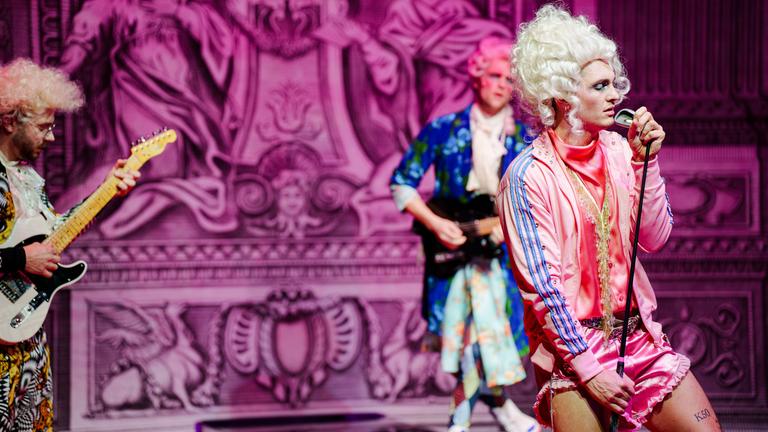 Bühnenszene: Eine Rockband, die Rokoko-Perücken und bunte Sportkleidung trägt spielt vor barocker Kulisse.