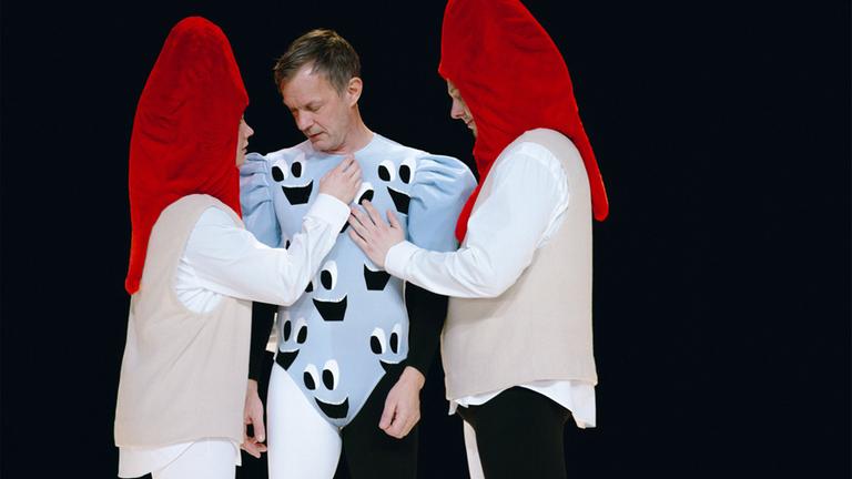 Bühnenszene: Ein Mann in hellblauem Turnanzug ist umgeben von zwei Personen, die rote Kopfbedeckungen tragen.