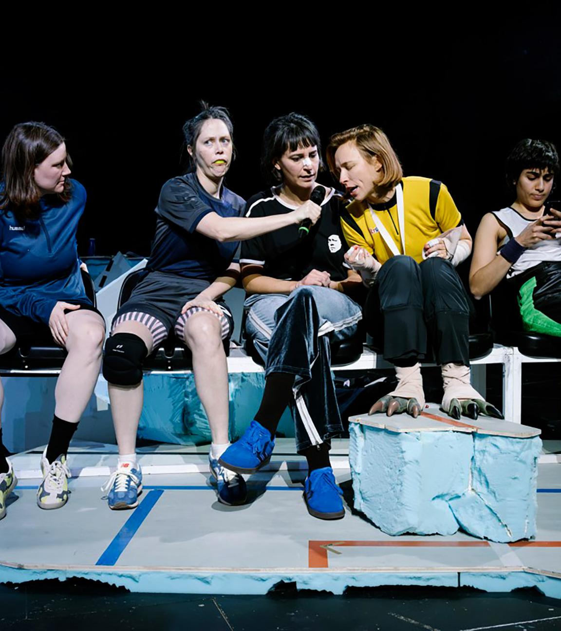 Bühnenszene: Frauen in Sportkleidung sitzen auf einer Bank.