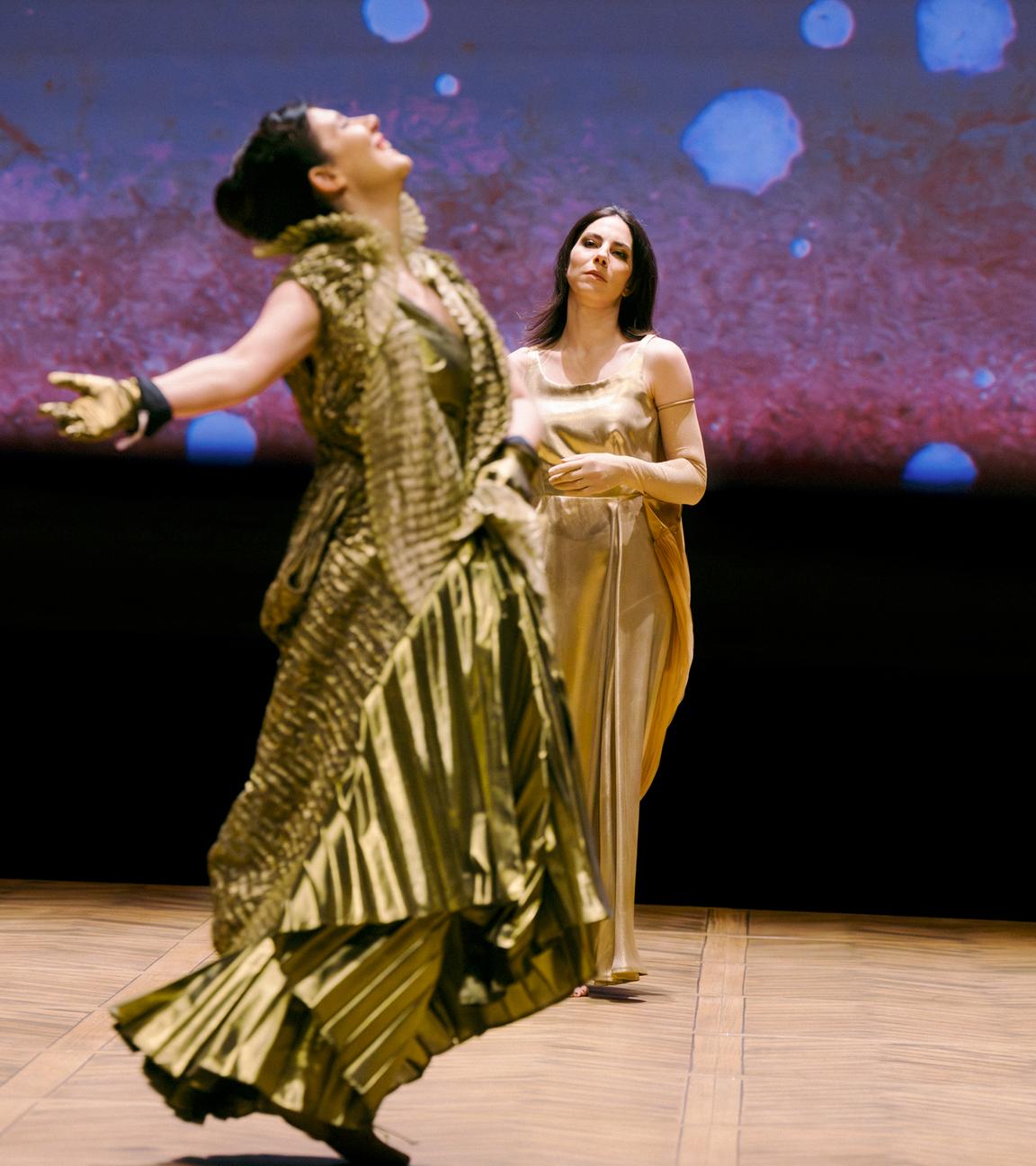 Bühnenszene: Zwei Frauen in goldenen Kleidern tanzen und stehen auf der Bühne.