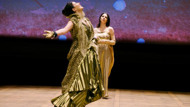 Bühnenszene: Zwei Frauen in goldenen Kleidern tanzen und stehen auf der Bühne.