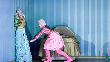 Bühnenszene: Zwei Frauen in bunten, retro-futuristischen Kostümen stehen vor einem Bett.