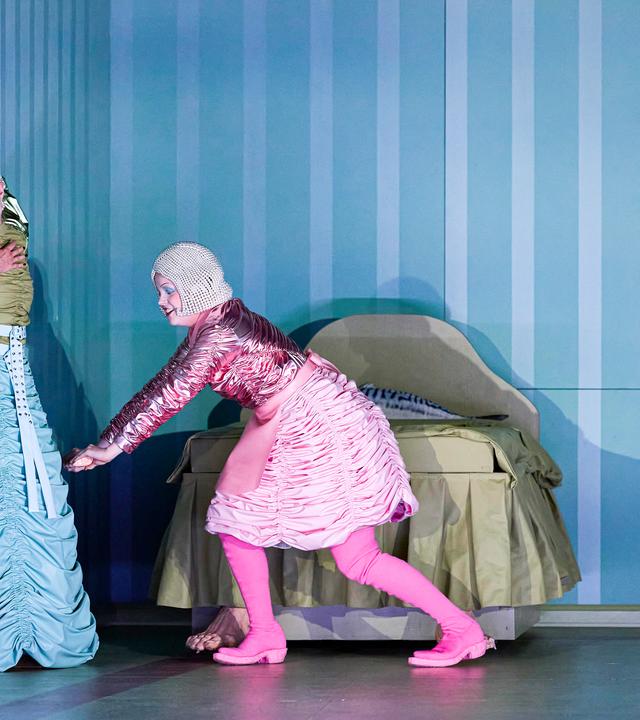 Bühnenszene: Zwei Frauen in bunten, retro-futuristischen Kostümen stehen vor einem Bett.