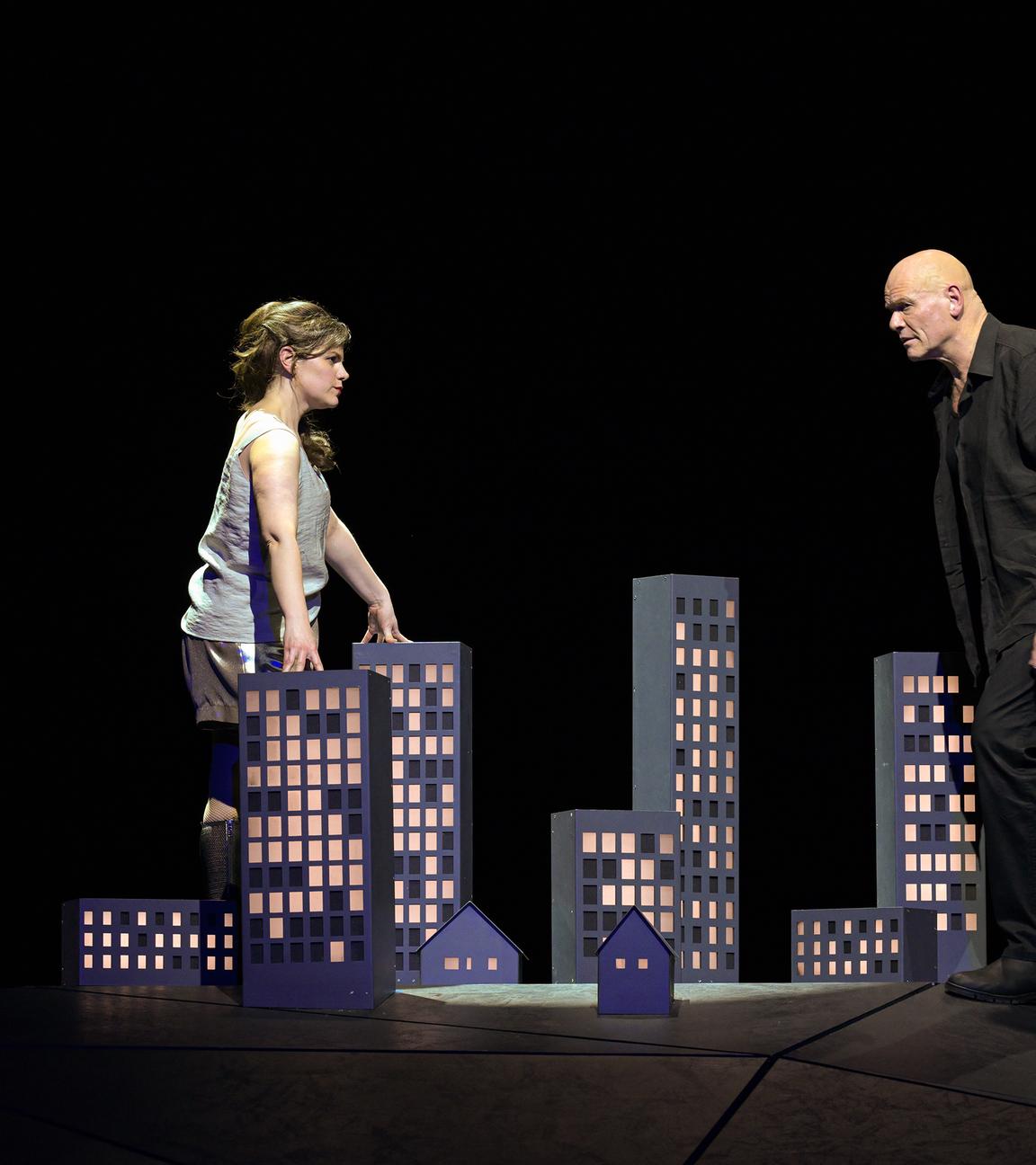 Bühnenszene: Eine Frau und ein Mann stehen sich gegenüber und sehen sich an. Zwischen ihnen steht auf dem Boden das Modell einer Stadt.