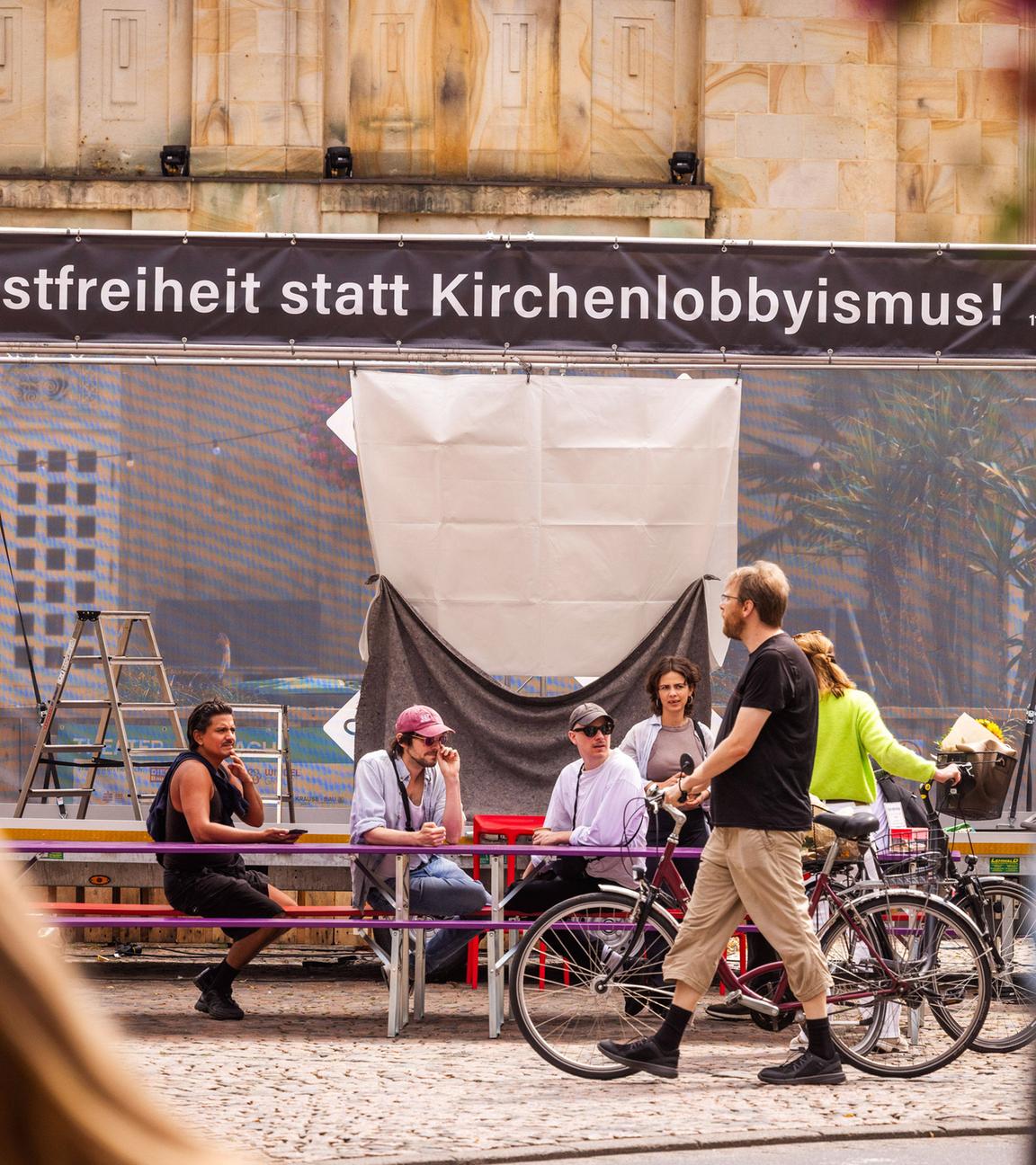 Vor einem großen Gebäude ist eine Bühne aufgebaut, über der ein Banner mit der Aufschrift Kunstfreiheit statt Kirchenlobbyismus steht. Davor sitzen ein paar Menschen auf Bierbänken, andere laufen daran vorbei.