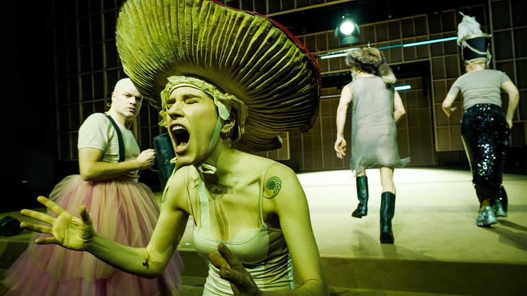 Bühnenszene: Eine Frau mit einem Pilzhut schreit, links daneben steht ein Mann, der ein rosa Tütü trägt. Zwei kostümierte Personen befinden sich im Hintergrund.