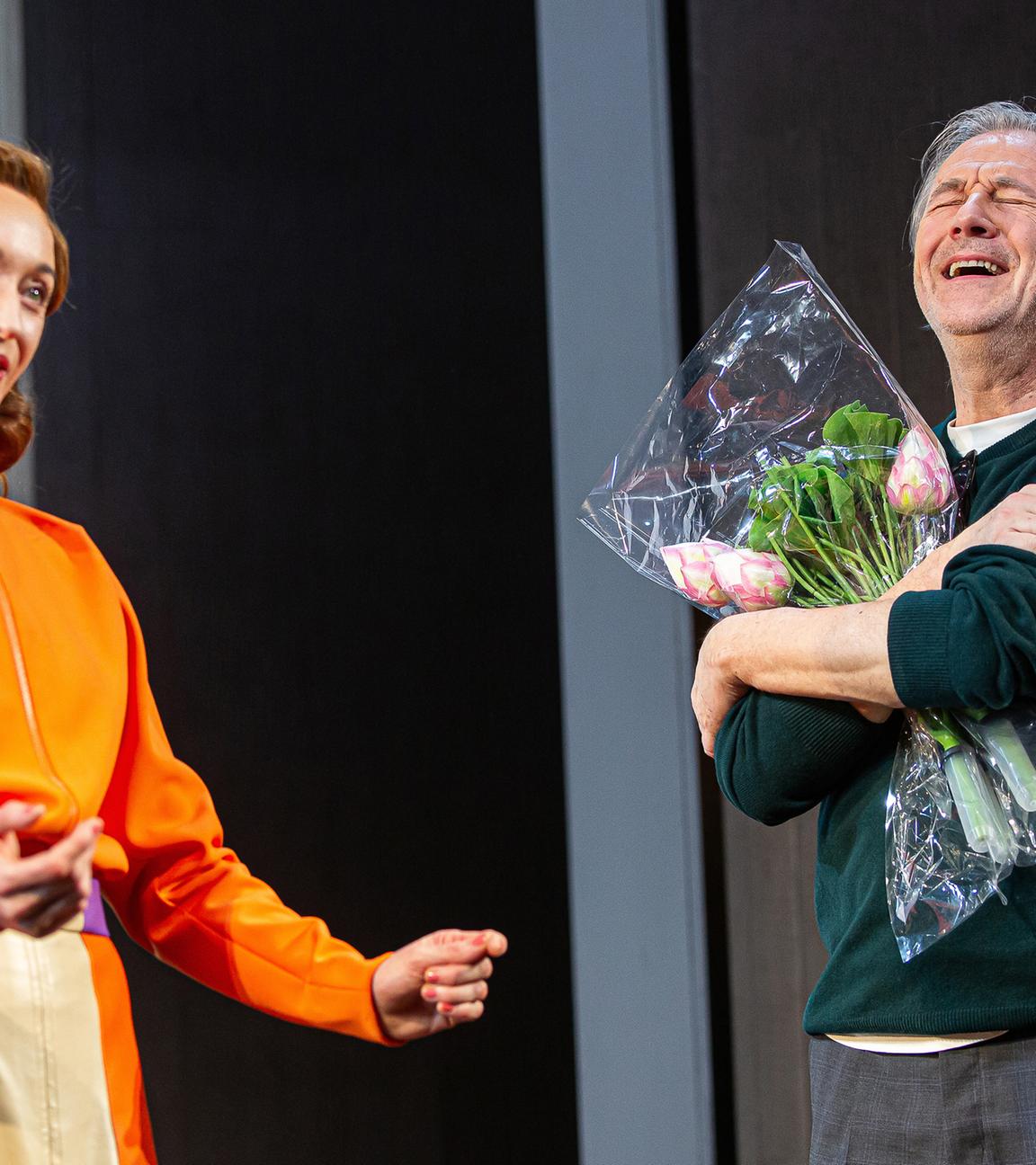 Bühnenszene: Eine Frau in orange-farbenem Kleid steht neben einem Mann, der einen Blumenstrauß in den Armen hält.