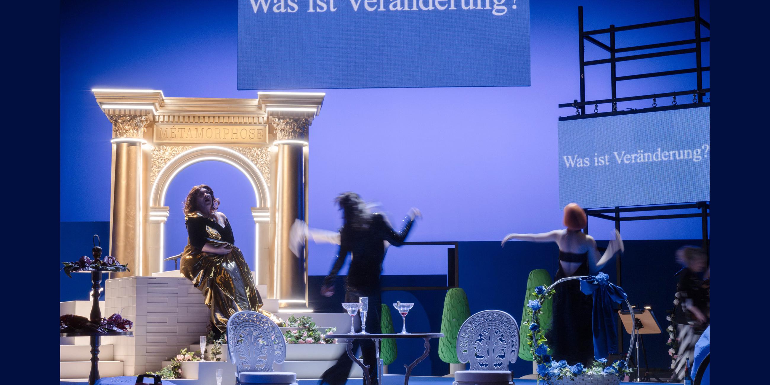 Bühnenszene: Drei Frauen sitzen und tanzen in einem Wohnzimmer. Im Hintergrund ist ein Portal zu sehen. Oben hängt ein Schild mit einer Aufschrift. Die Bühne ist blau angeleuchtet.