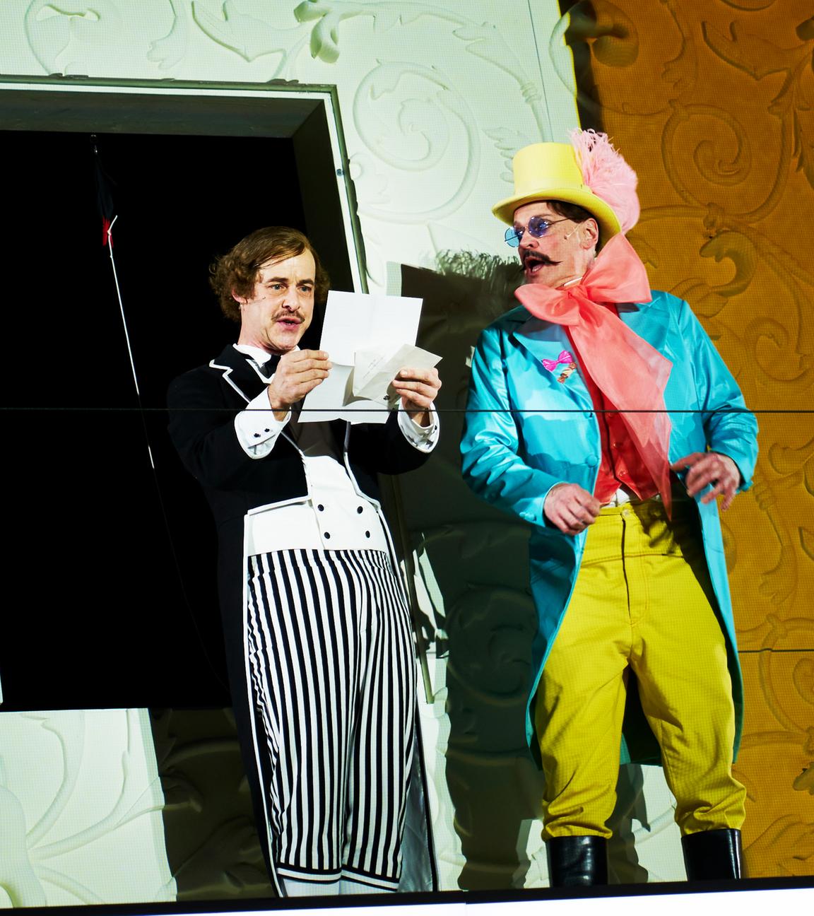 Bühnenszene: Ein Mann im Frack liest einen Brief, ein anderer Mann, der einen bunten Frack und Zylinder trägt, schaut ihn an.